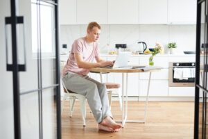 Young casual man sitting by table in the kitchen in front of laptop